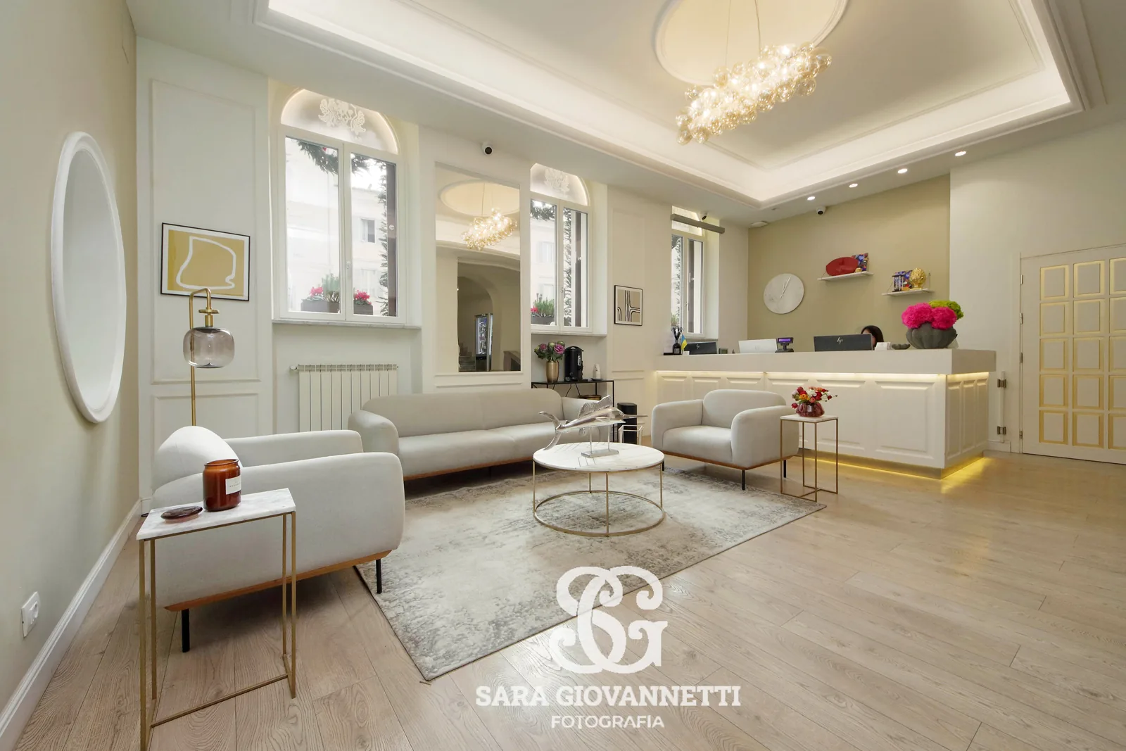Fotografo Airbnb Roma: hall elegante con divani chiari, lampadario di cristallo, bancone reception illuminato e fiori colorat