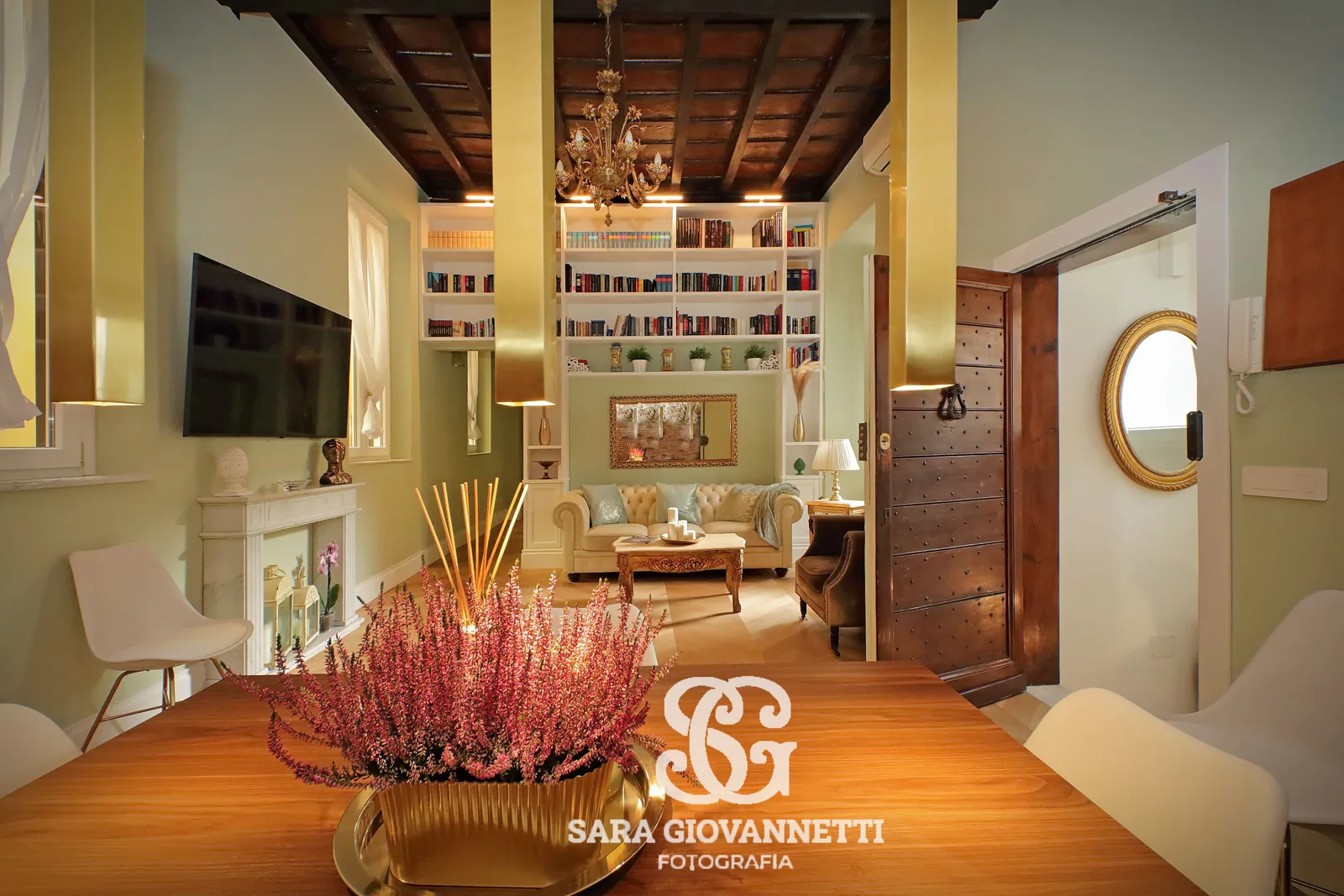 Fotografo Airbnb Roma riprende elegante soggiorno con soffitto a cassettoni, libreria a muro, divano Chesterfield e fiori ros