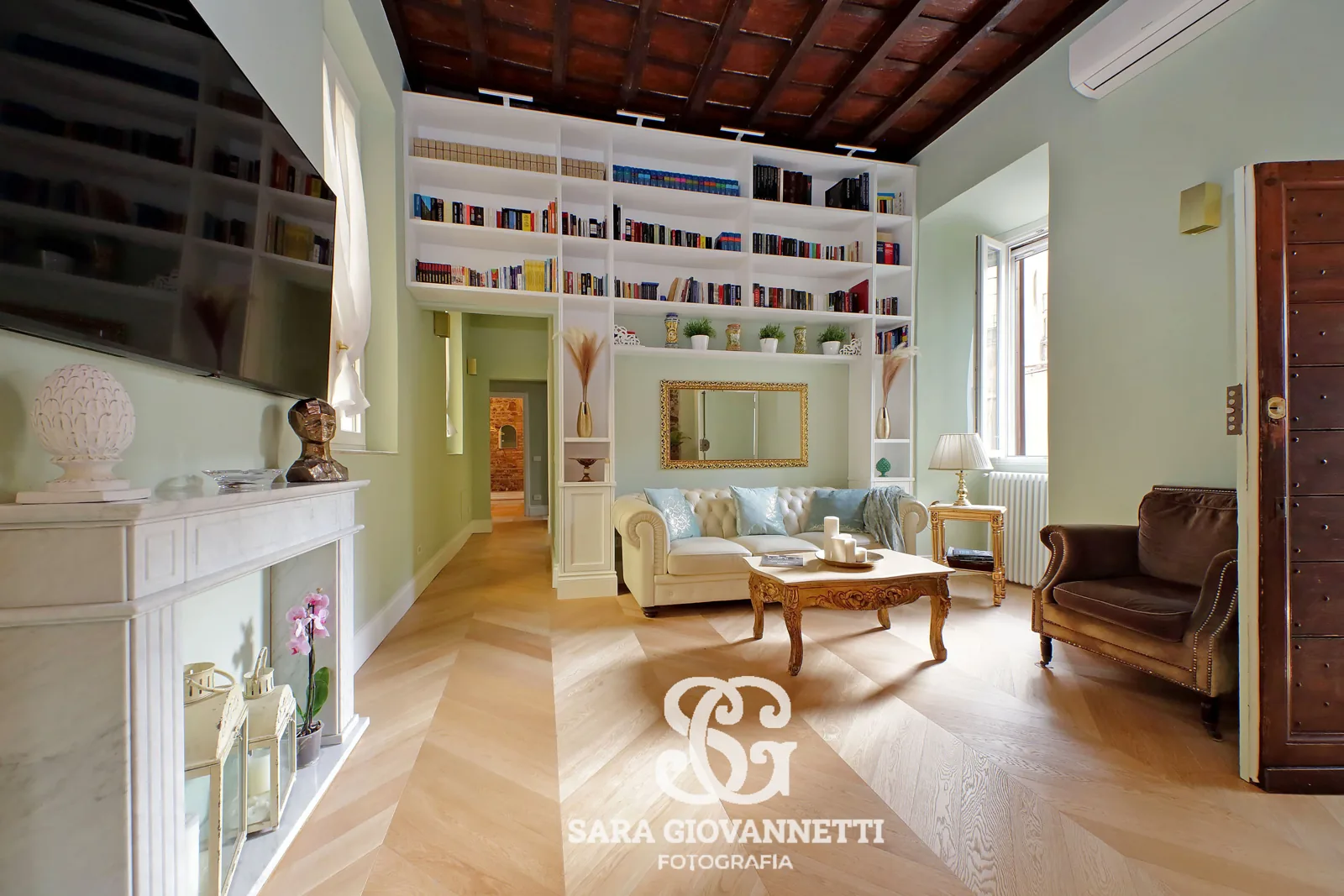 Fotografo Airbnb Roma ritrae elegante soggiorno con libreria a tutta parete, soffitto a cassettoni e camino in marmo